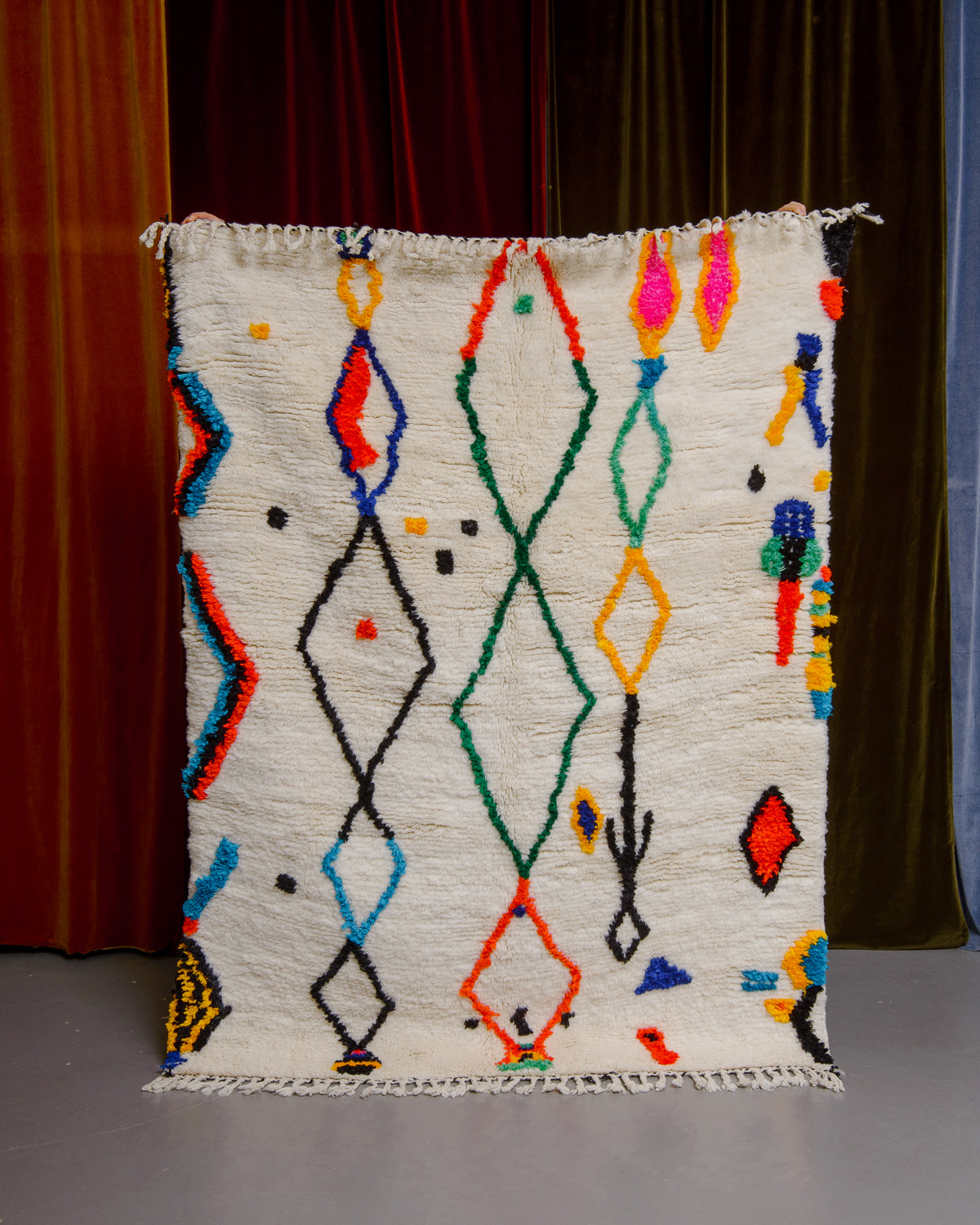 Berberlin Finest rug with a thick cream pile and colourful design in cotton being held in front of velvet curtains. 
