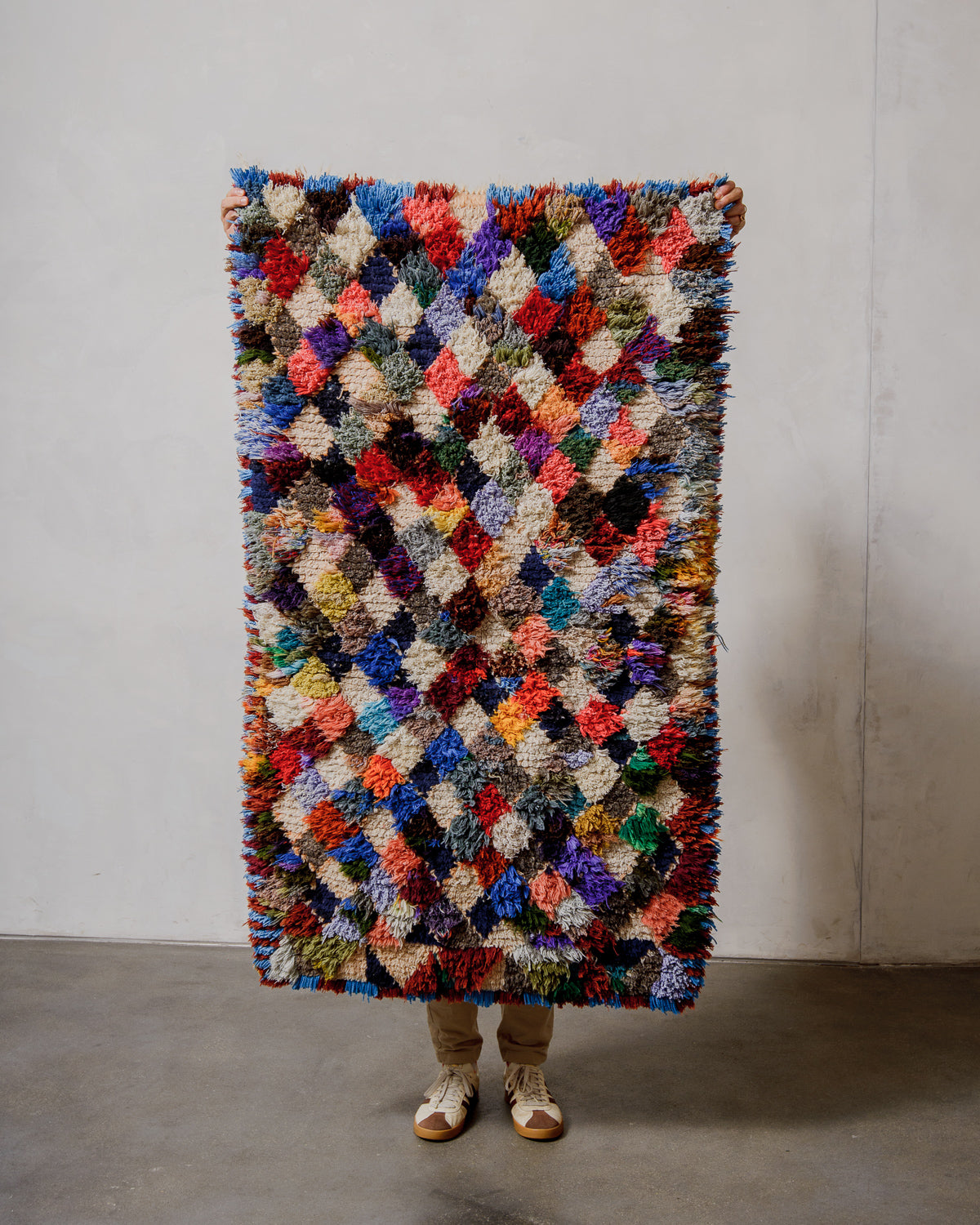 Multicoloured textured rug held by a person against a plain wall.