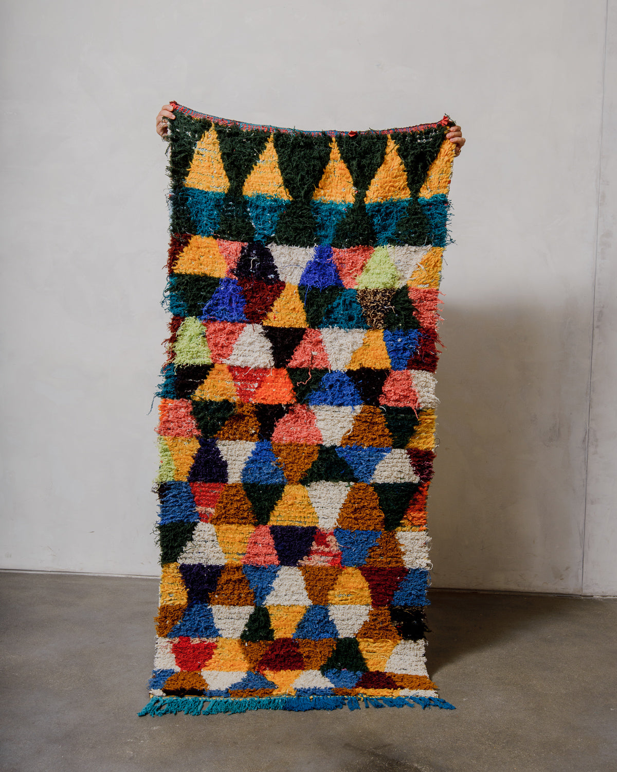 Multicoloured geometric patterned rug held by a person against a plain background.