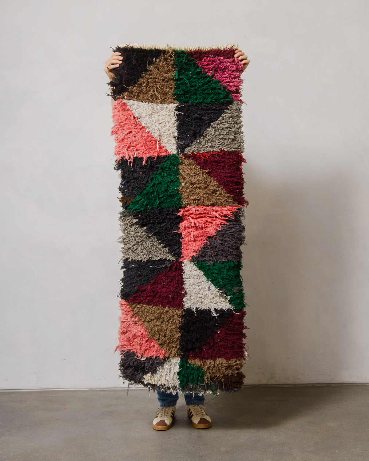 Person holding a colorful geometric-patterned rug against a plain wall.