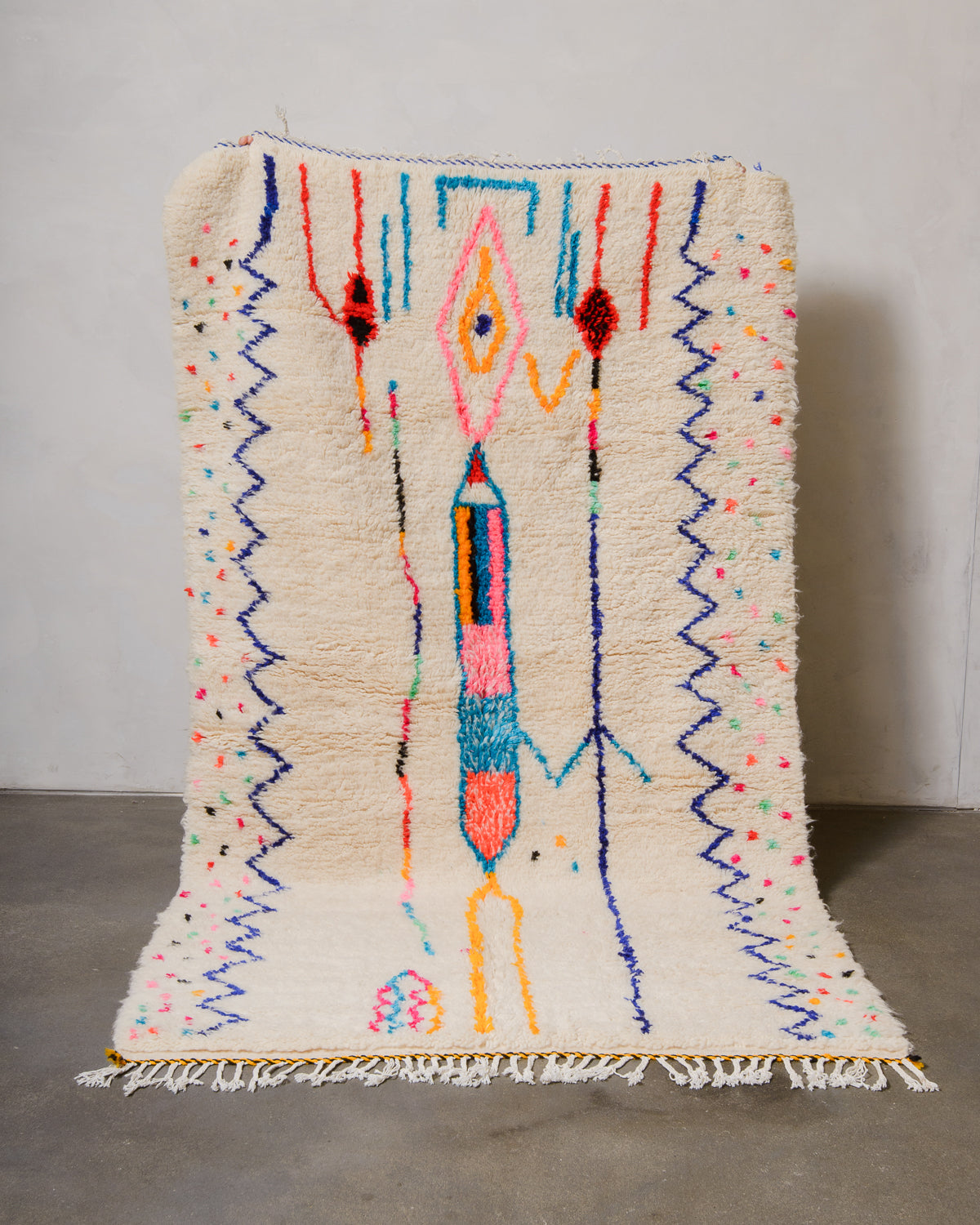 Berberlin Azilal rug made of fluffy cream wool and colourful design, held up in front of a white wall and cement floor.