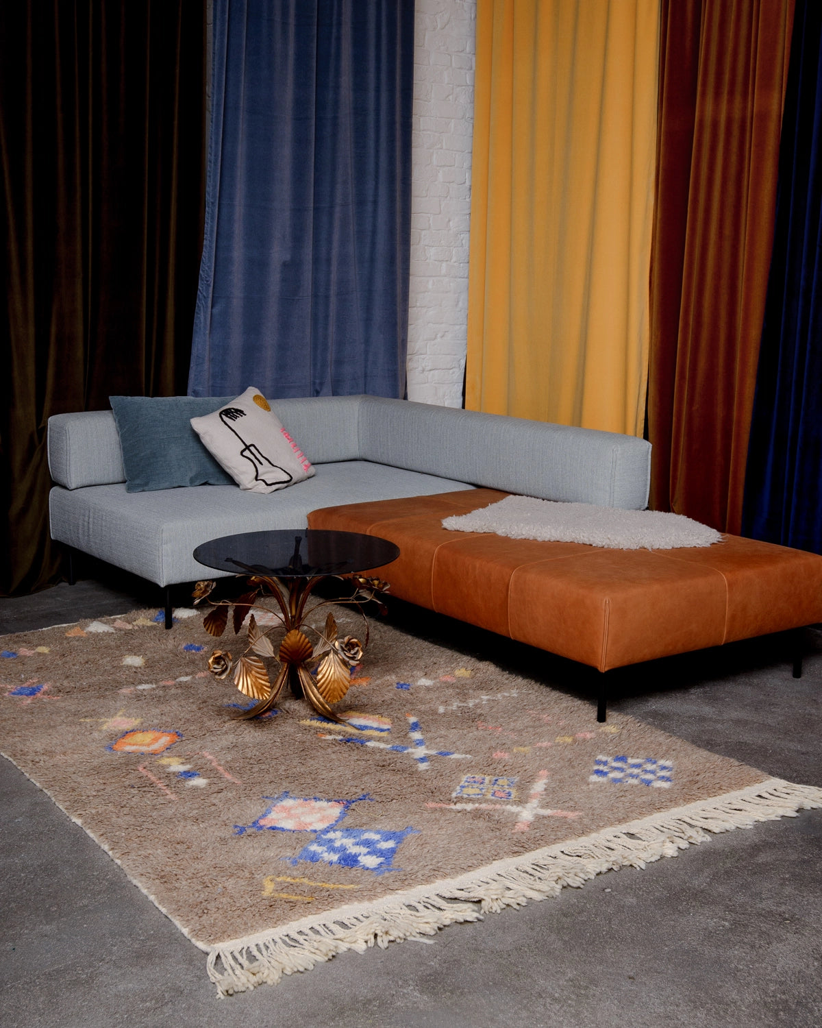 Berberlin vintage boujad rug in soft brown wool with symbols in blue, pink, yellow and white, laying under modular couch, with different coloured velvet curtains behind.