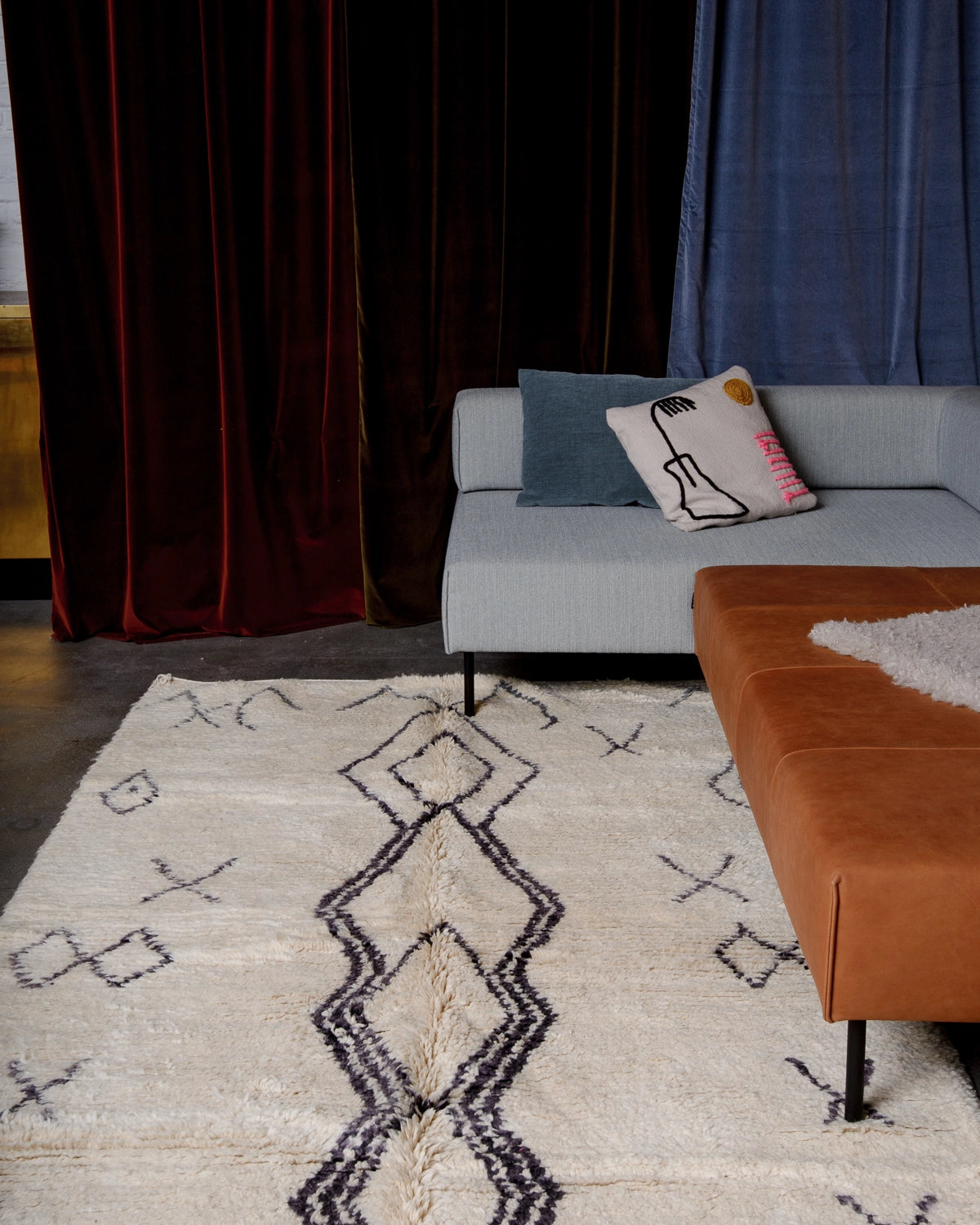 Berberlin vintage boujad rug in cream wool with dark grey symbols laying under modern sectional couch, with different coloured velvet curtains behind.