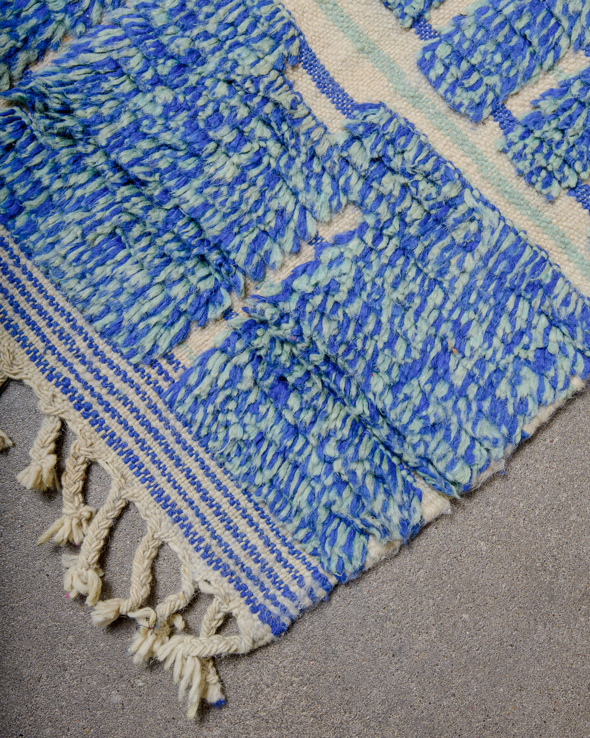 Corner of blue and turquoise berber rug with striped woven end and natural wool tassels.