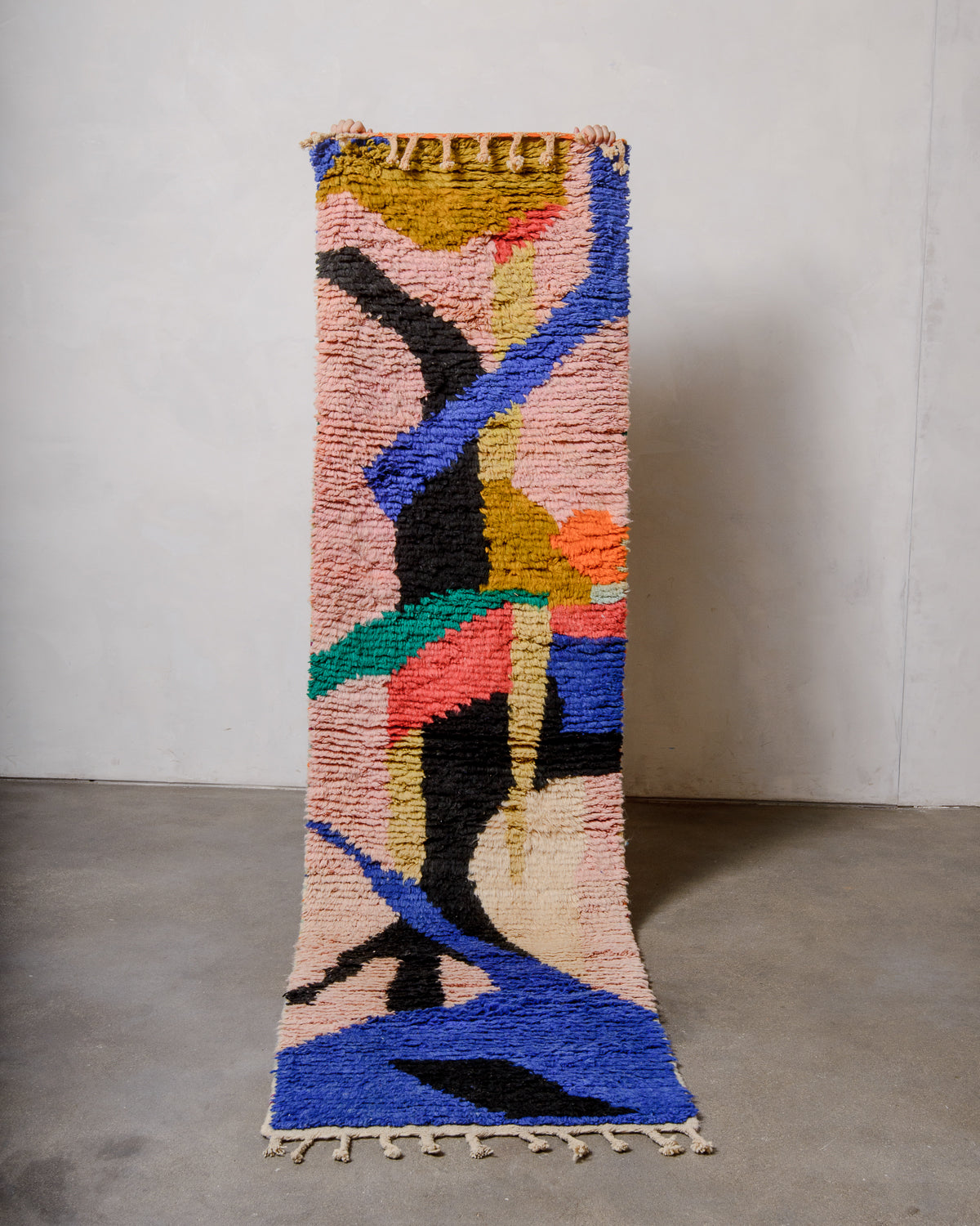 Berberlin vintage runner rug made of brightly coloured wool, being held up in front of a white wall and concrete floor. 