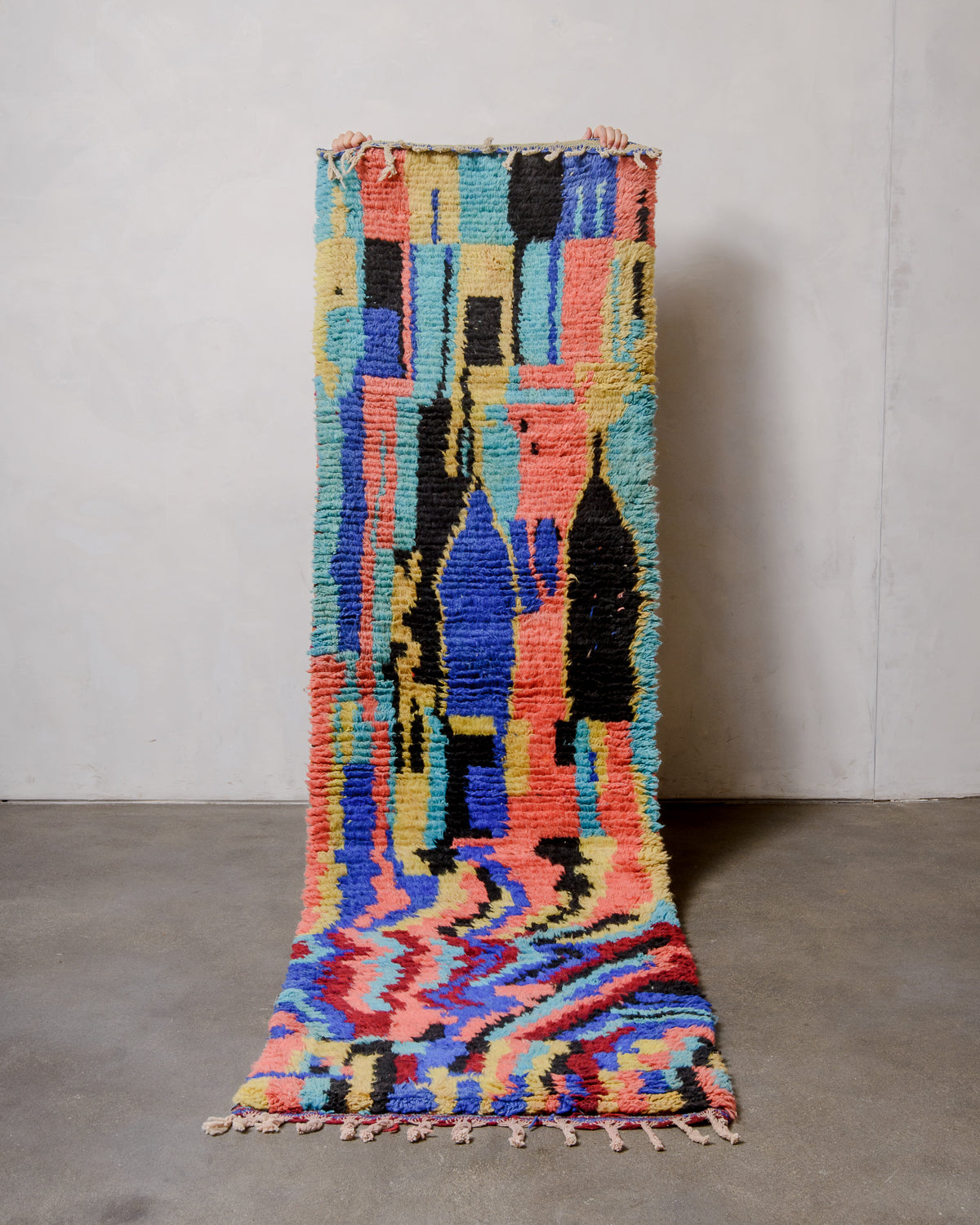 Berberlin vintage runner rug made of brightly coloured wool, being held up in front of a white wall and concrete floor. 