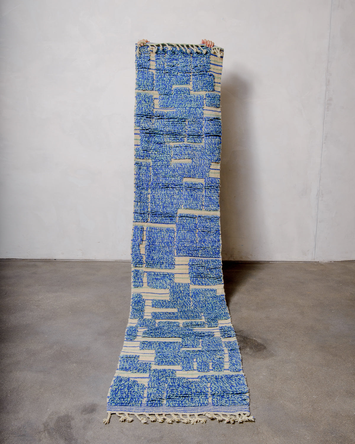 Berberlin vintage Berber runner rug made of blue and turquoise wool, being held up in front of a white wall and concrete floor. 