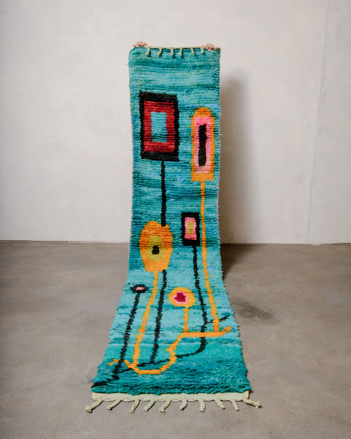 Berberlin vintage Berber runner rug made of colourful wool, being held up in front of a white wall and concrete floor. 