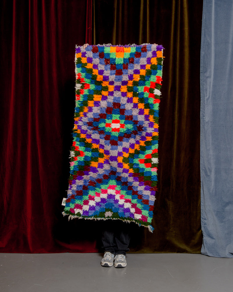 Colourful boucherouite vintage rug with a diamond pattern made up of small squares being held in front of velvet curtains.
