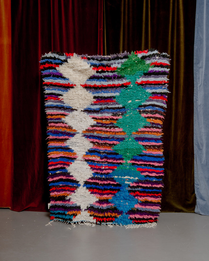 Vintage Berberlin boucherouite runner rug being held up in front of a velvet curtain. 