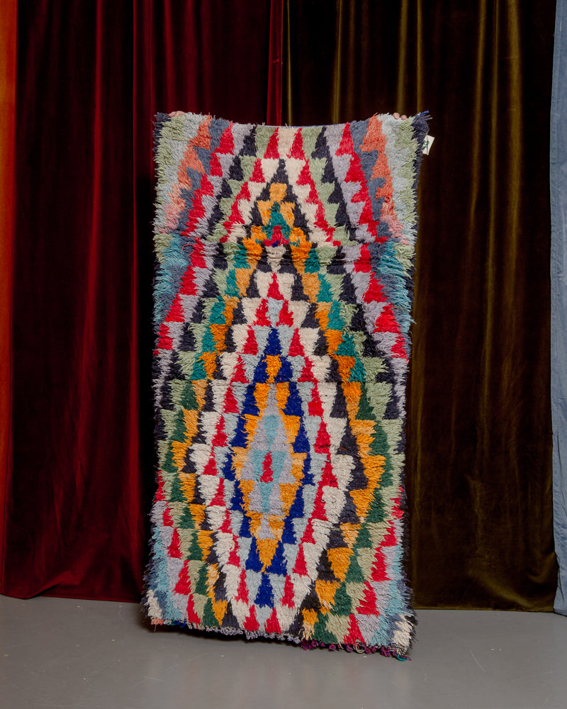 Vintage Berberlin boucherouite runner rug being held up in front of a velvet curtain. 
