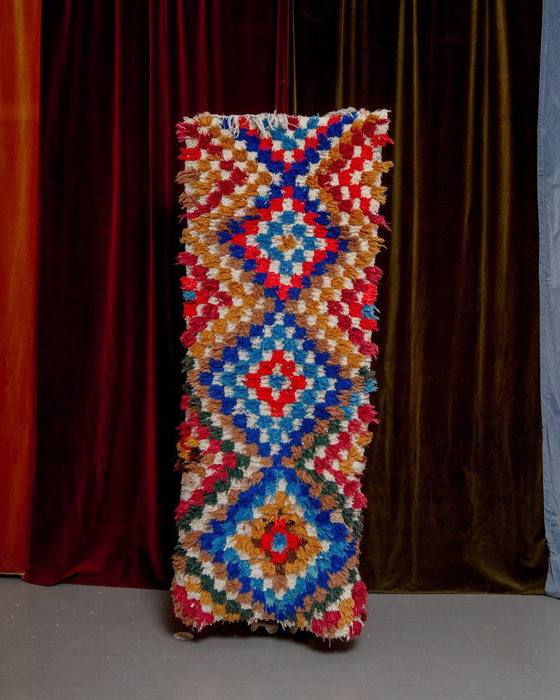 Vintage Berberlin boucherouite runner rug being held up in front of a velvet curtain. 
