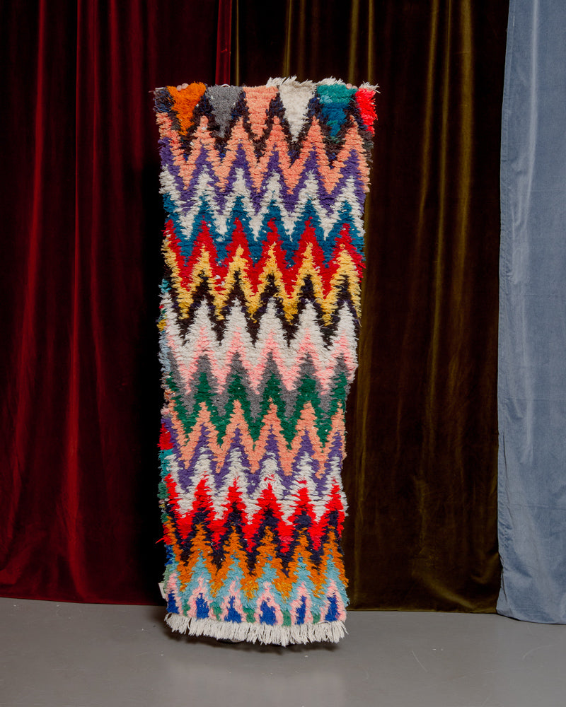 Vintage Berberlin boucherouite runner rug being held up in front of a velvet curtain. 