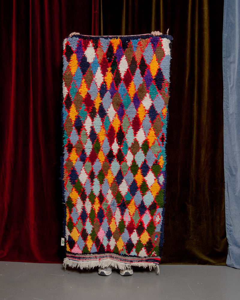 Vintage Berberlin boucherouite runner rug being held up in front of a velvet curtain. 