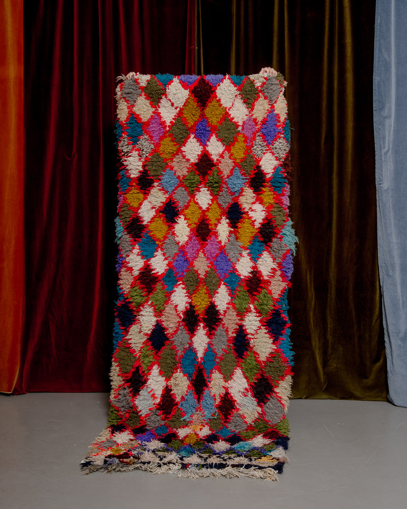 Vintage Berberlin boucherouite runner rug being held up in front of a velvet curtain. 