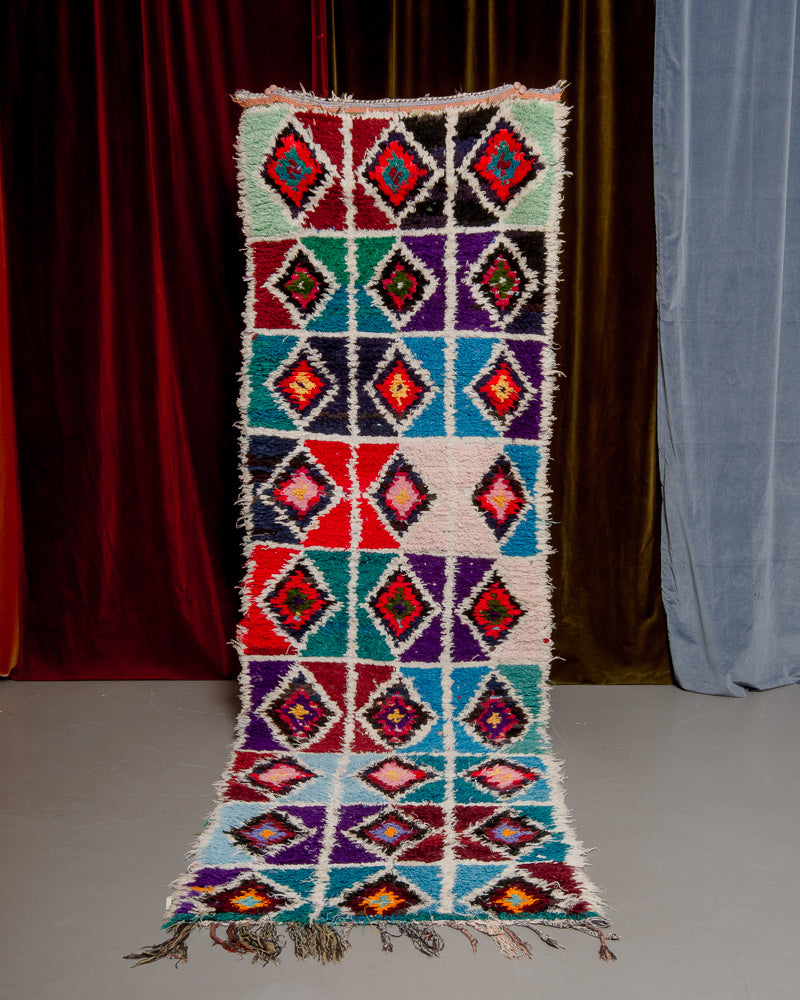 Vintage Berberlin boucherouite runner rug being held up in front of a velvet curtain. 