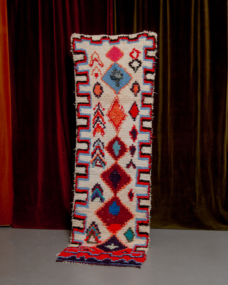 Vintage Berberlin boucherouite runner rug being held up in front of a velvet curtain. 
