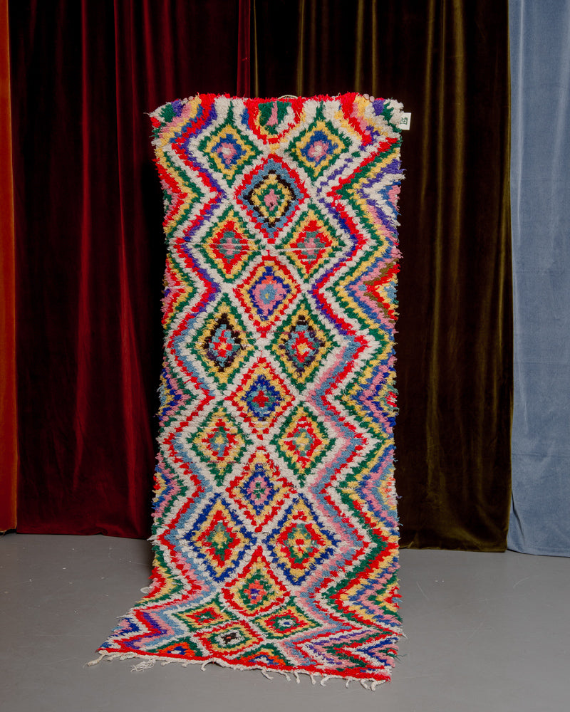 Vintage Berberlin boucherouite runner rug being held up in front of a velvet curtain. 