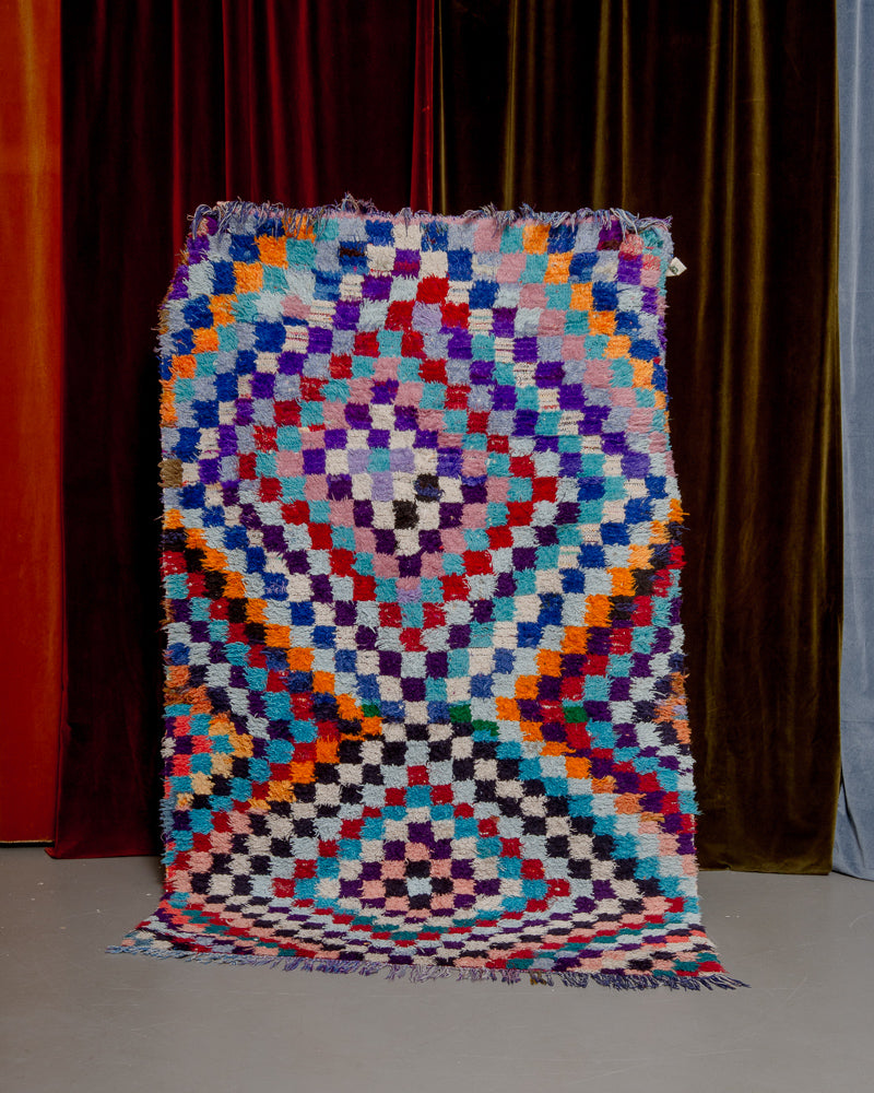 Colourful boucherouite vintage rug with a diamond pattern made up of small squares being held in front of velvet curtains.