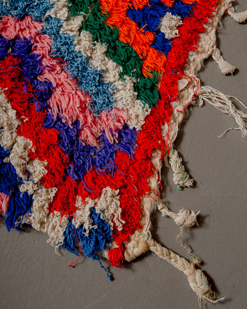 Corner of boucherouite rug with red, blue green and white yarn and tassels made of a mix of wool and plastic.