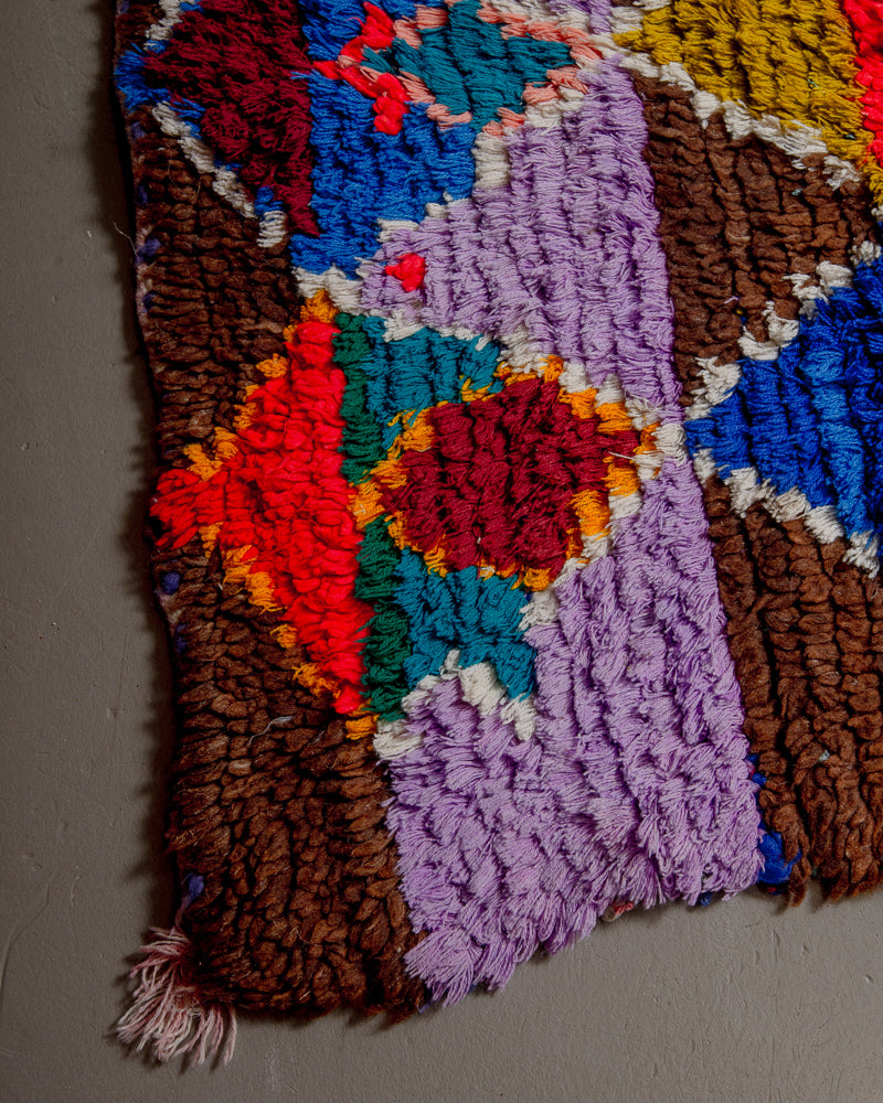 Corner of boucherouite rug with red, blue green and lila yarn and brown wool.