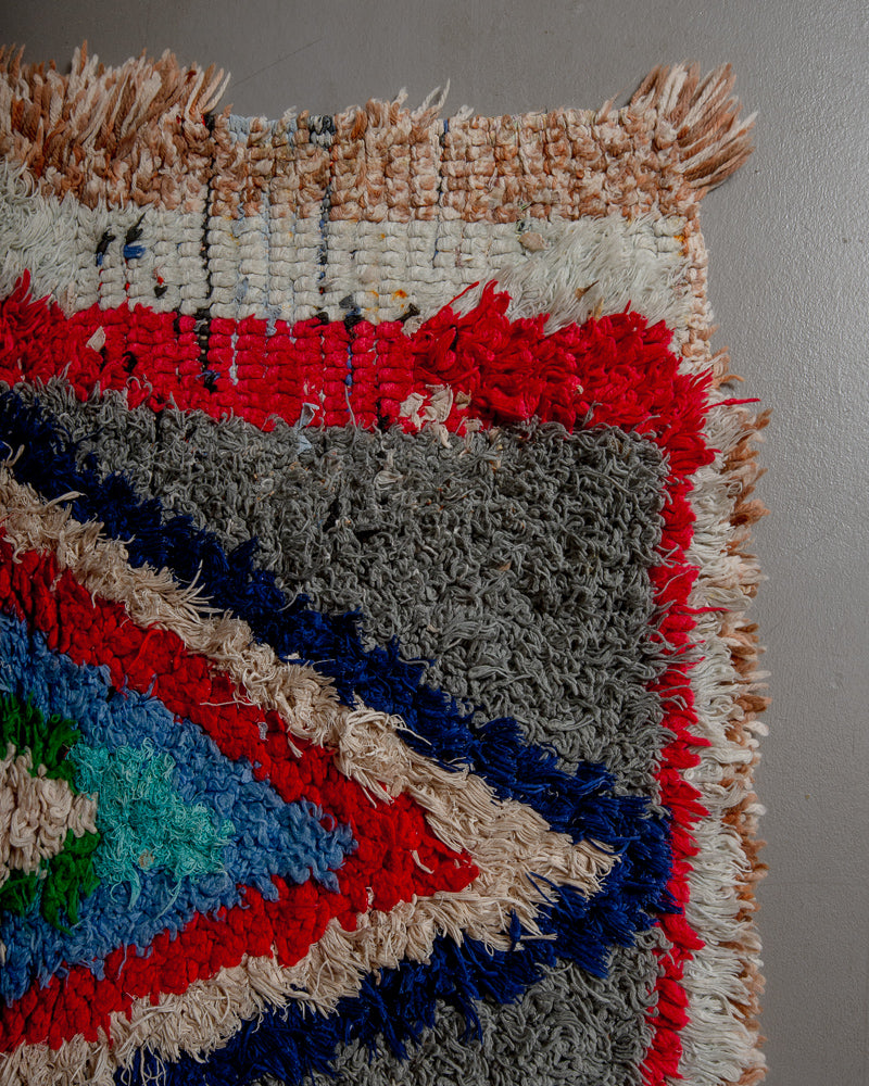 Corner of boucherouite rug with red, blue, grey and cream yarn.