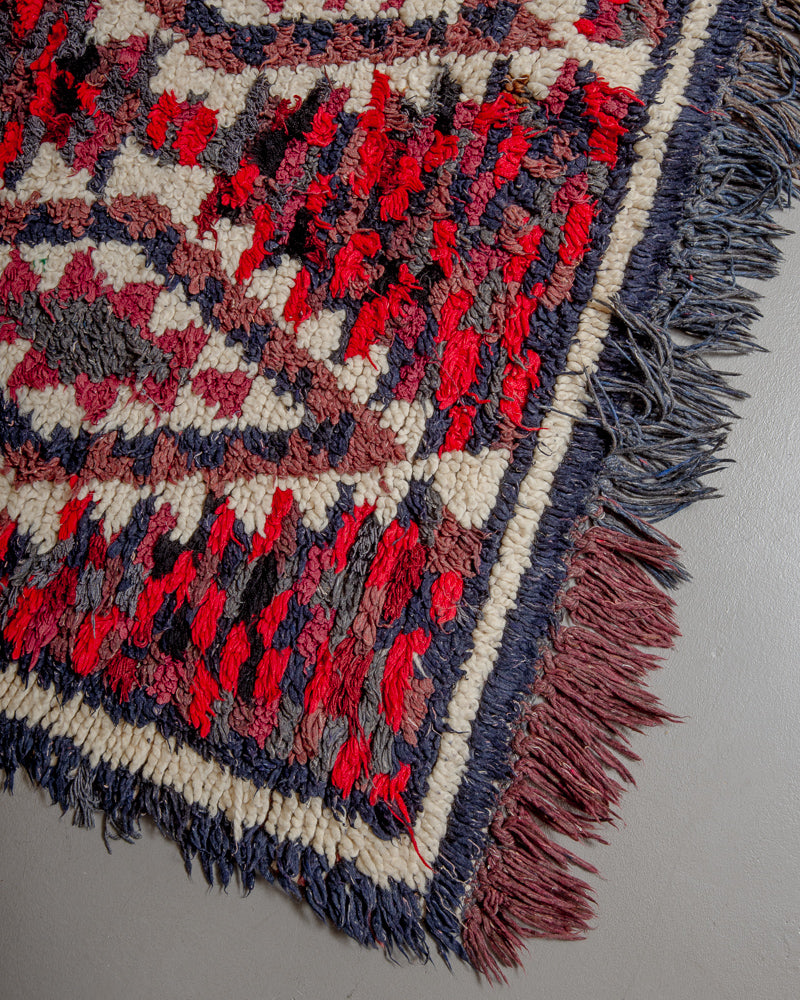 Corner of boucherouite rug with red, blue and brown yarn and cream wool and fringes along the edge.